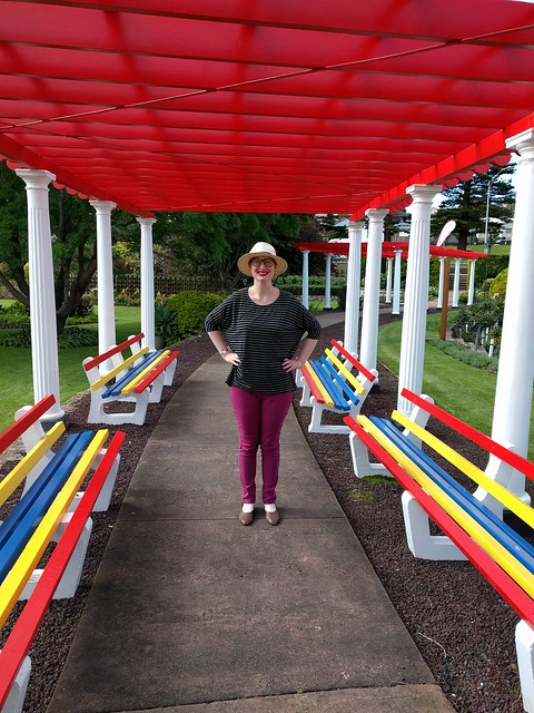 A woman stands under a pergola, wearing a draped knit tee, purple pants, and a panama hat.