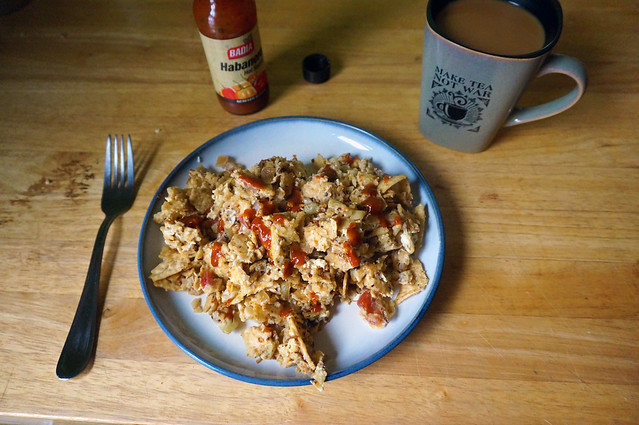 A plate of migas dotted with hot sauce, carefully arranged with a fork to the side, a mug of coffee to the upper right, and a bottle of hot sauce to the upper left.
