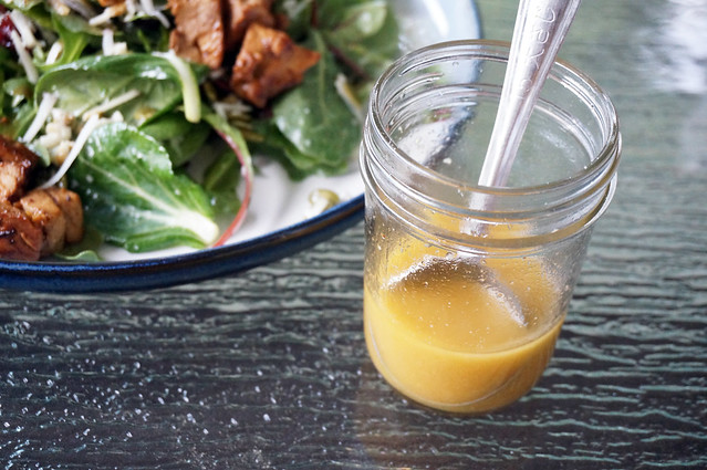 Golden yellow chicken vinaigrette in a small glass jar, resting near the edge of a plated of salad on the glass tabletop. On the bright metal tablespoon resting in the liquid, some stamped Japanese characters are discernable.