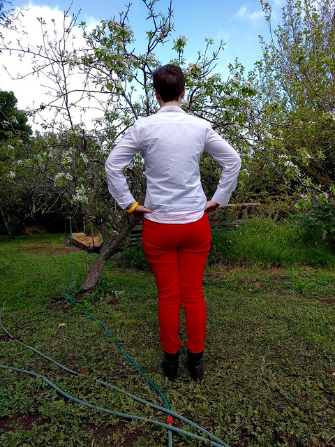 A woman stands in a garden, wearing a white button up shirt and red skinny pants.